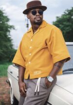 Black Man wearing a yellow KhayaKhulu Box Shirt, with a brown fedora hat and brown pants