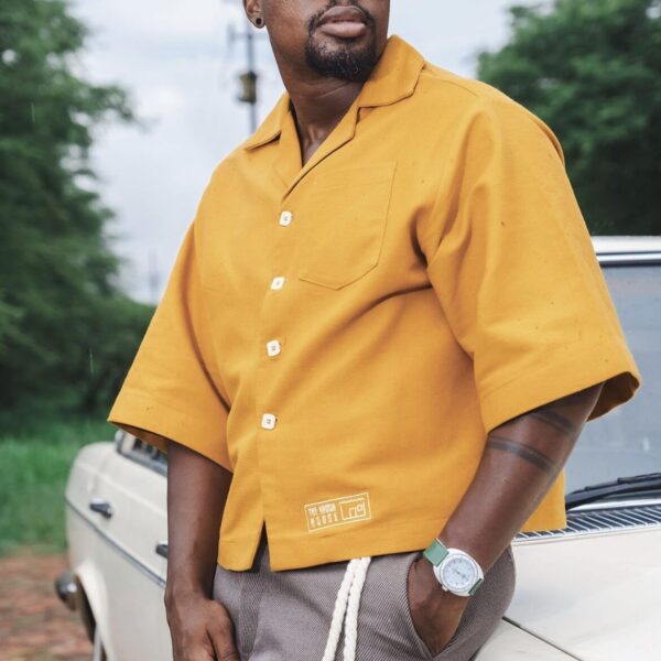 Black Man wearing a yellow KhayaKhulu Box Shirt, with a brown fedora hat and brown pants